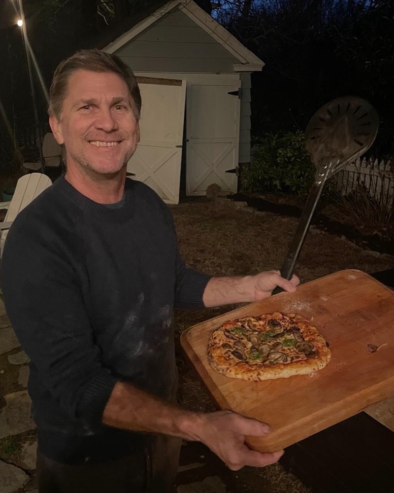 Patrick Noble in his backyard in the evening, holding a homemade pizza on a wooden board